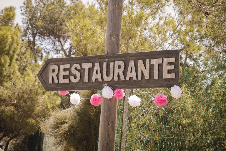 A rustic wooden restaurant sign in Spain, adorned with festive pink and white decorations amidst a natural outdoor setting.